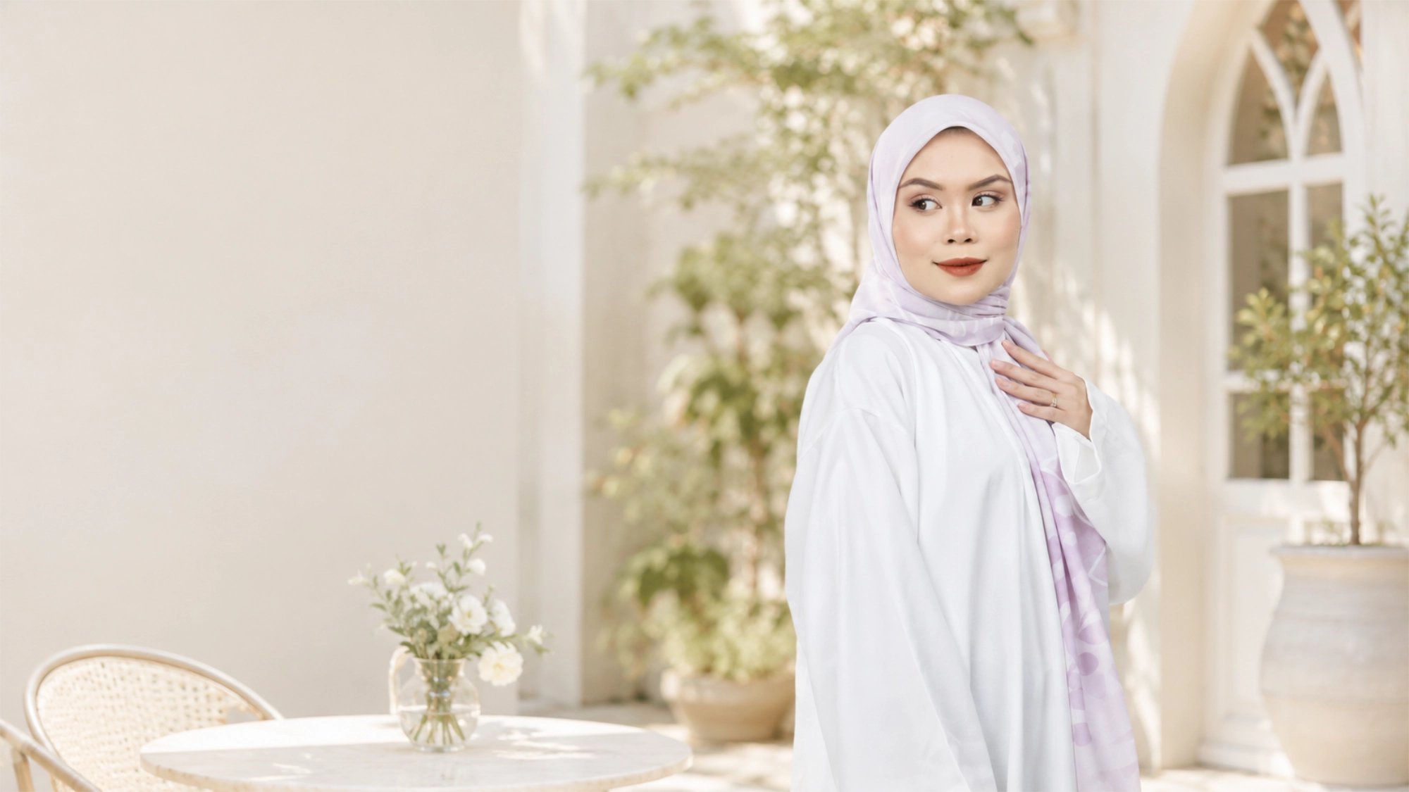 Model wearing a Tudung Bawal Cotton Voile Thistle by Hana Malaysia in a bright, indoor setting with plants and a table.
